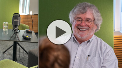 Wissenschaftler David Kaplan sitzt in einem Interviewraum. Neben ihm sind ein Mikrofon und Aufnahmegeräte zu sehen; ein eingeblendetes Wiedergabesymbol kennzeichnet das Bild als Video.
