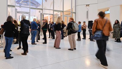 Menschen stehen in kleinen Gruppen in einem hellen, großzügigen Veranstaltungsraum und unterhalten sich. Im Vordergrund geht eine Person durch den Raum.