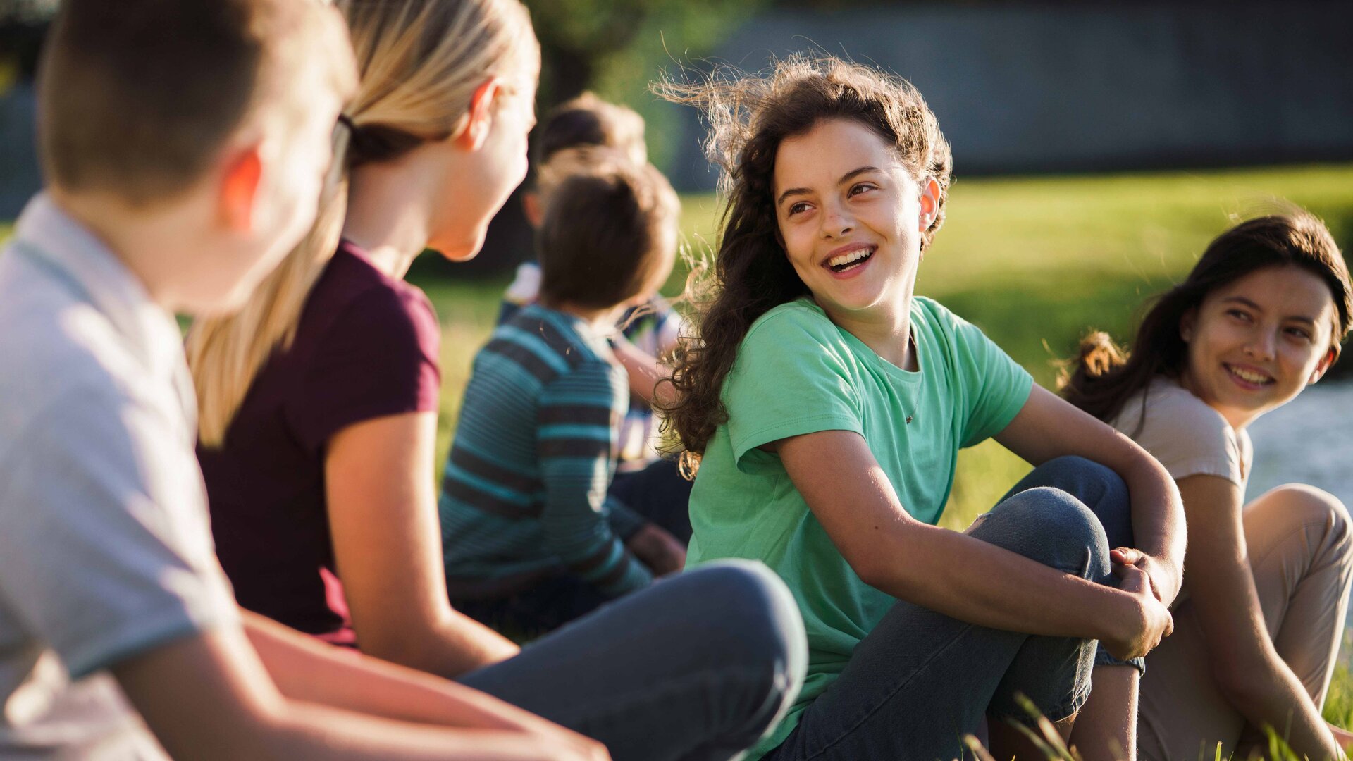 Eine Gruppe von Kindern sitzt im Gras. Zwei Mädchen schauen lachend die anderen an